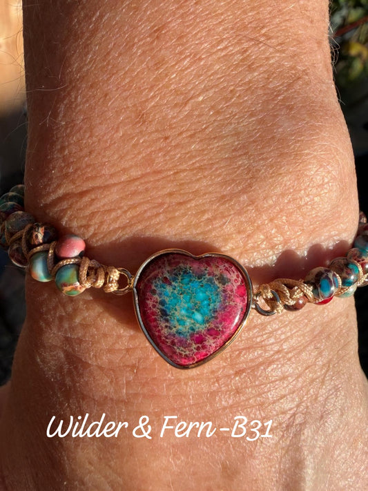 A close-up of a person's wrist wearing a handmade Boho beaded bracelet with a heart-shaped turquoise and pink stone centerpiece and gold-toned metal links. The text 'Wilder & Fern-B31' is overlaid at the bottom. By Free Spirit
