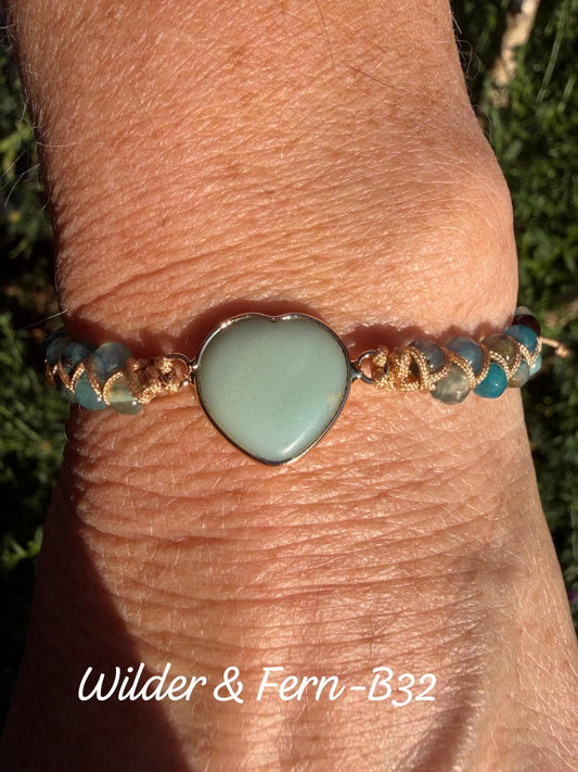 A close-up of a person's wrist wearing a handmade bracelet. The bracelet features a central heart-shaped light blue-green stone surrounded by gold-toned metal and small blue-green beads on either side By Free Spirit